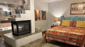 A bedroom with a fireplace, a bed with patterned bedding, and artwork on the walls. - Riverside Retreat (Ruidoso)