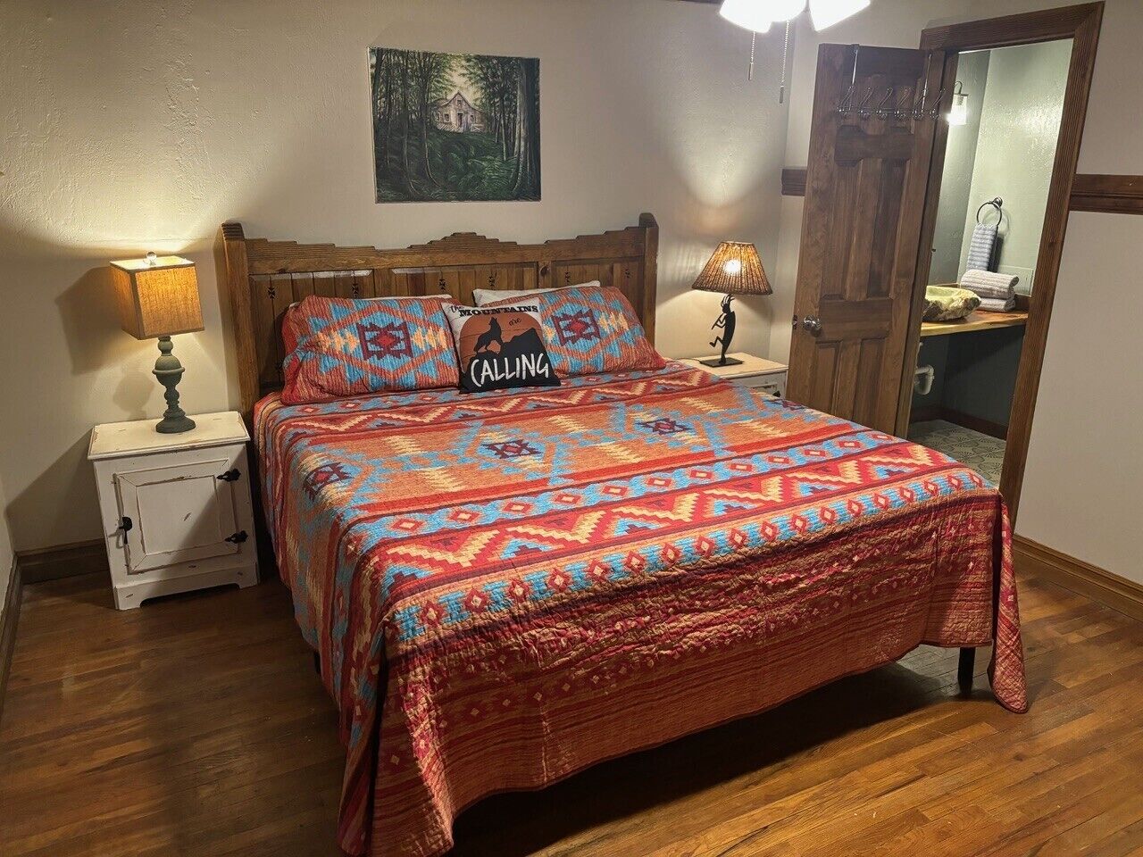 Een slaapkamer met een houten bed, beddengoed met een patroon, een nachtkastje met een lamp, een klein wit kastje en een schilderij aan de muur.