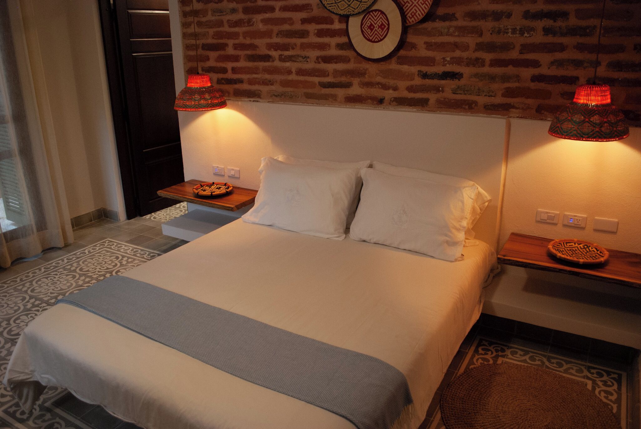 A neatly made bed with white linens, two bedside tables with decorative plates, and a brick wall with hanging wall art.