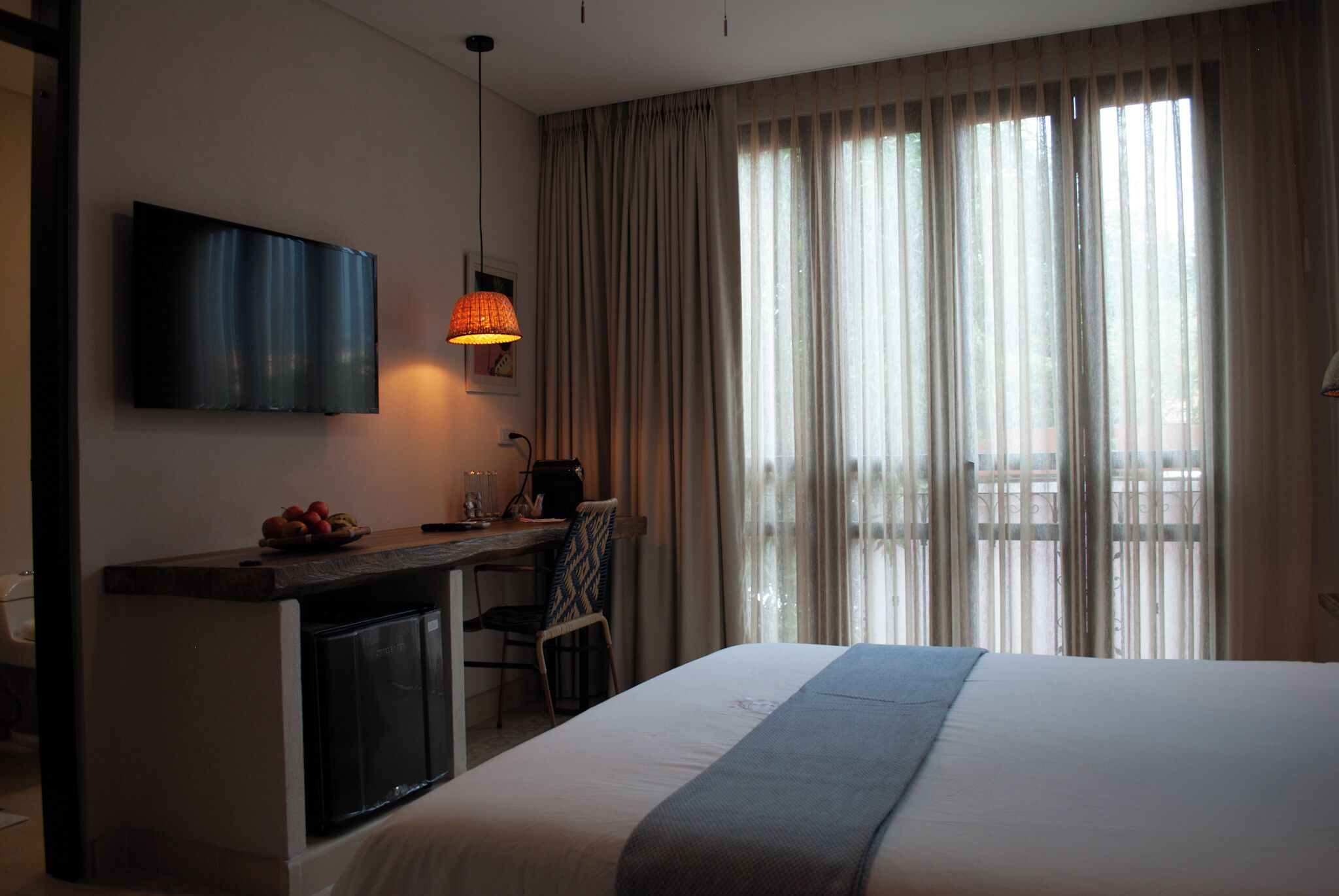 A hotel room with a bed, a desk with a computer, a TV, and a view of the outdoors through large windows.
