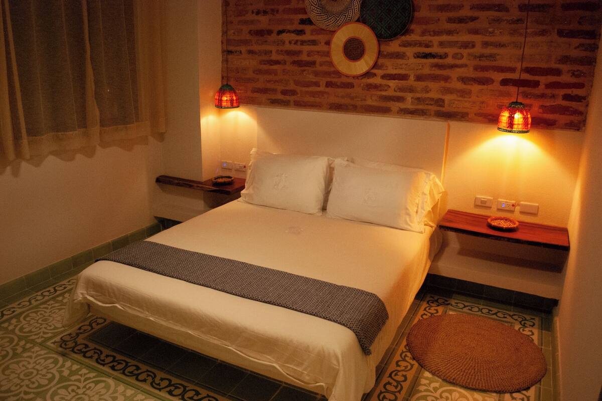 A neatly made bed with white linens, two bedside tables with lamps, a brick wall, and patterned floor tiles.