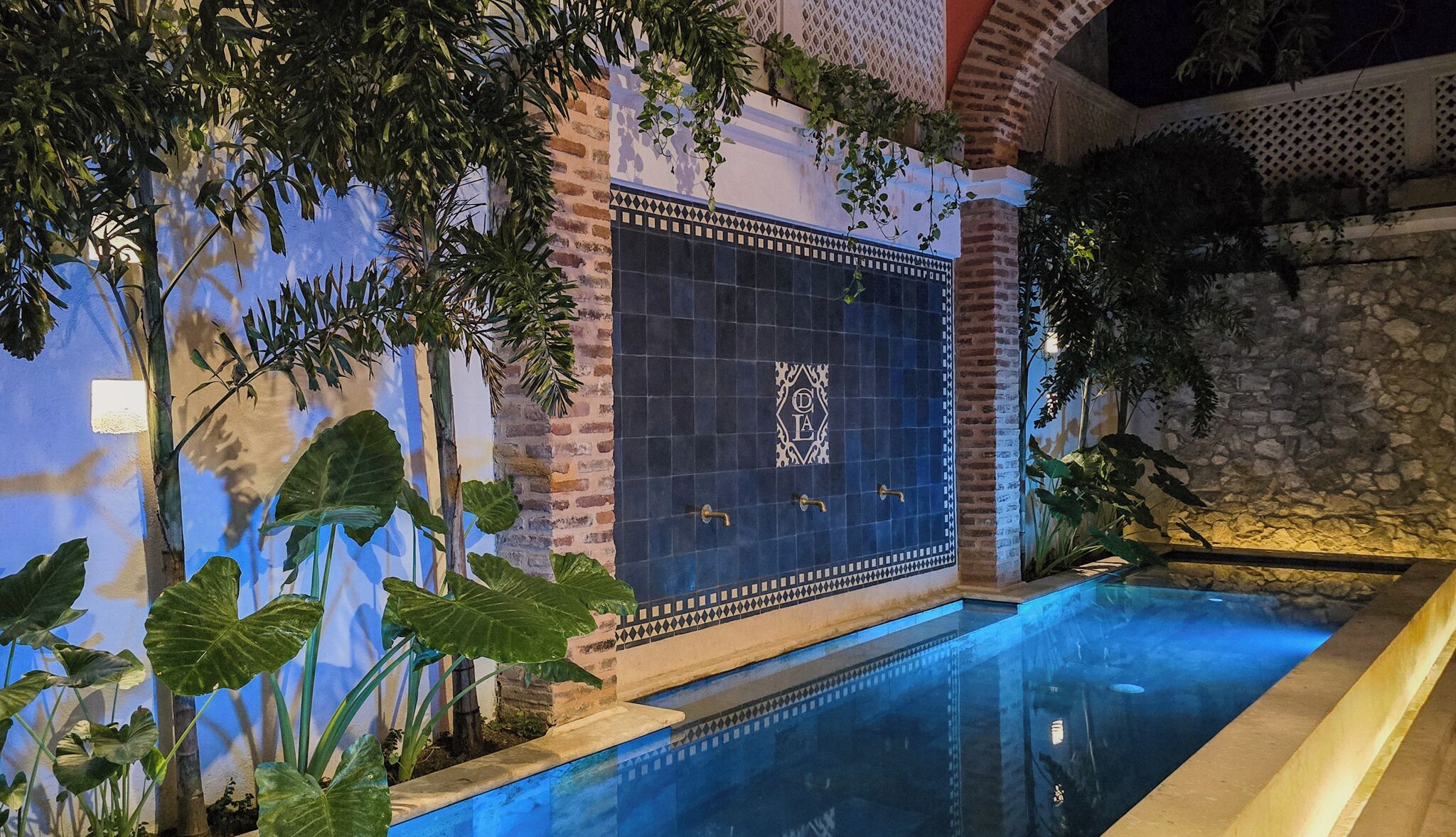 Un área de piscina con un muro de azulejos, un muro de piedra y grandes plantas verdes.