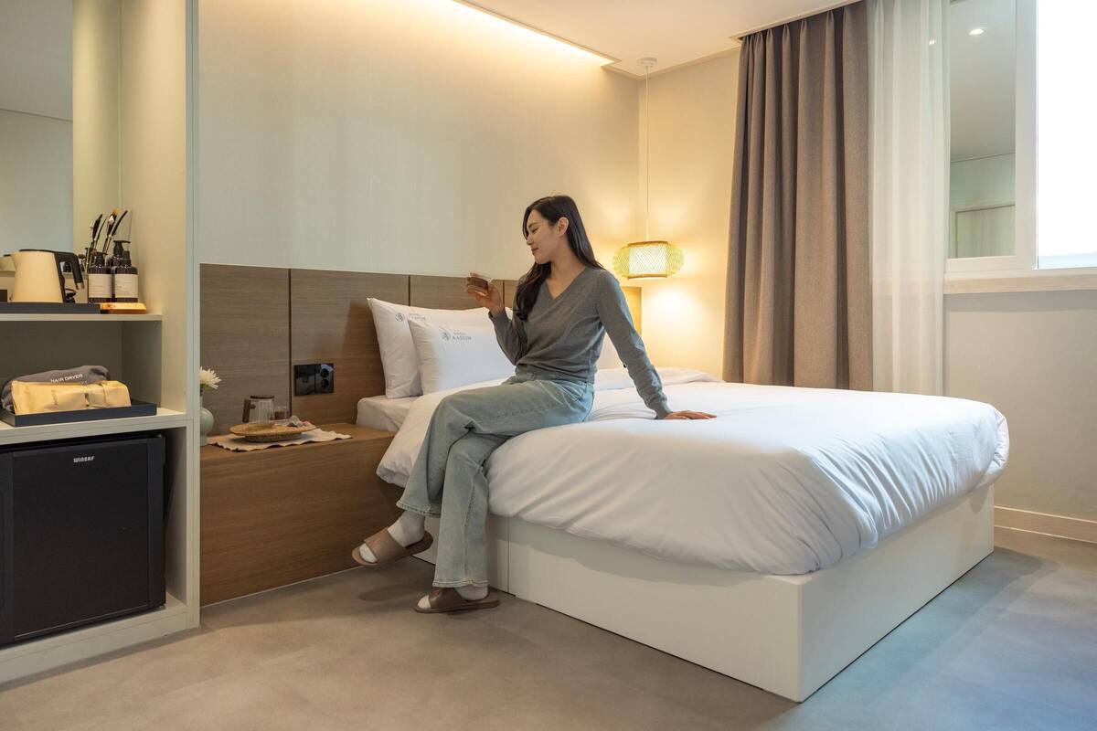Habitación de hotel con cama, mesita de noche con lámpara, un refrigerador pequeño y una ventana con cortinas.