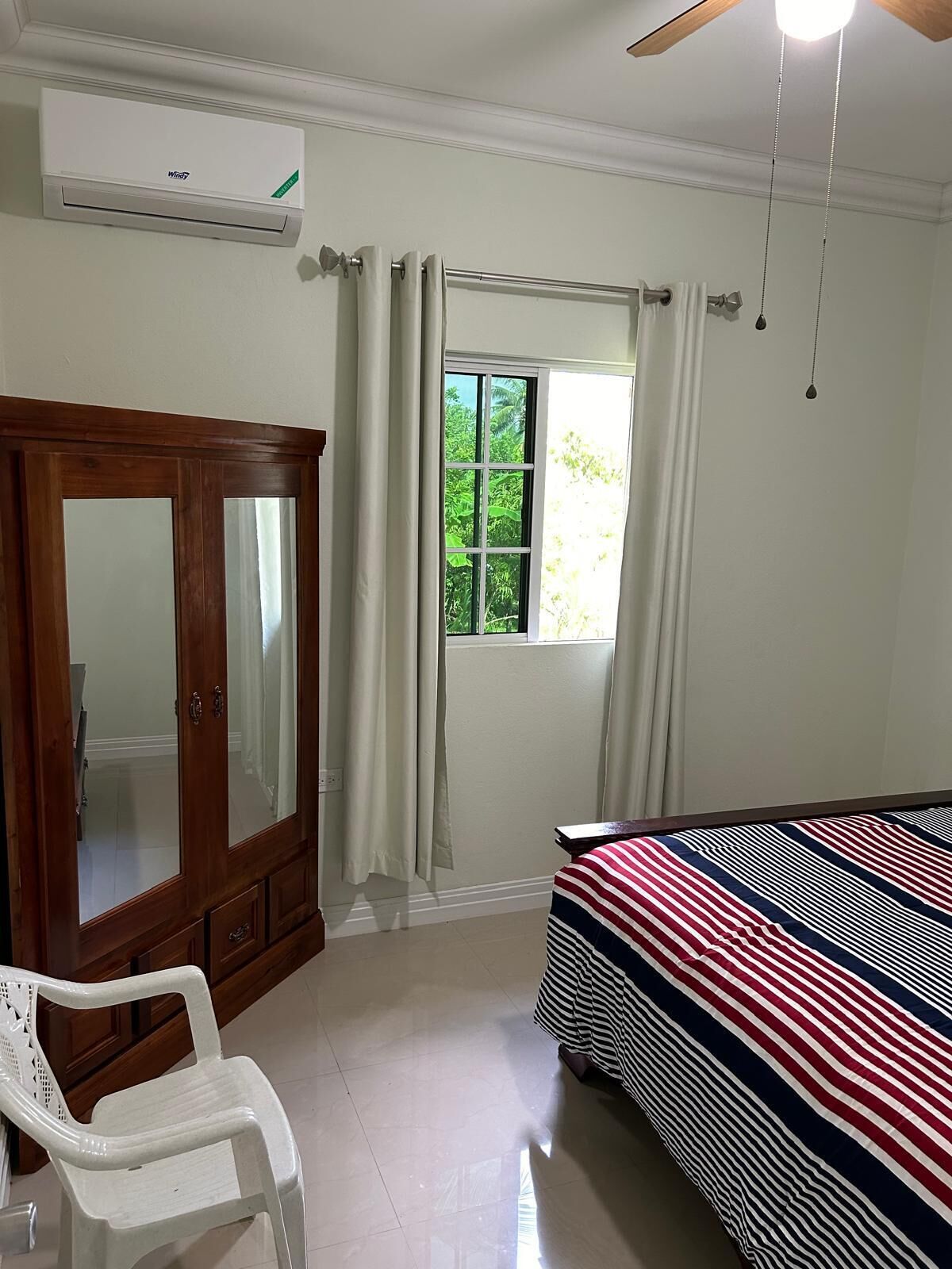 A bedroom with a bed, a wardrobe, a window with curtains, a ceiling fan, and a white air conditioner.