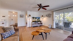 Un salón amplio con ventilador de techo, un ventanal con vistas a palmeras, una zona de comedor con mesas redondas y una acogedora sala de estar con sofá y cojines.