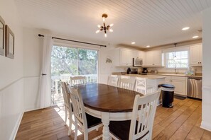 Un comedor con mesa de madera y sillas blancas, una cocina con muebles blancos y electrodomésticos de acero inoxidable, y un ventilador de techo.