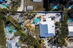 An aerial view of a residential area with multiple swimming pools, palm trees, and a large building with a sloped roof.