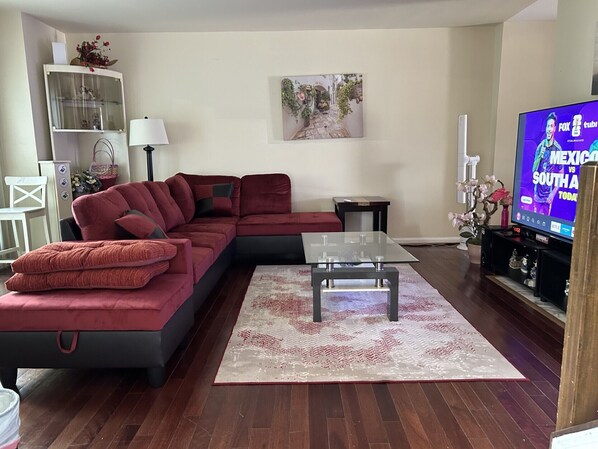A living room with a sectional sofa, a glass coffee table, a TV displaying a sports event, and a decorative wall piece.