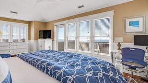 A bedroom with a large bed, a TV, a desk with a chair, and a window with blinds. - Discover your dream beach house with an amazing ocean view and lots of deck space on South Beach! (Bald Head Island)