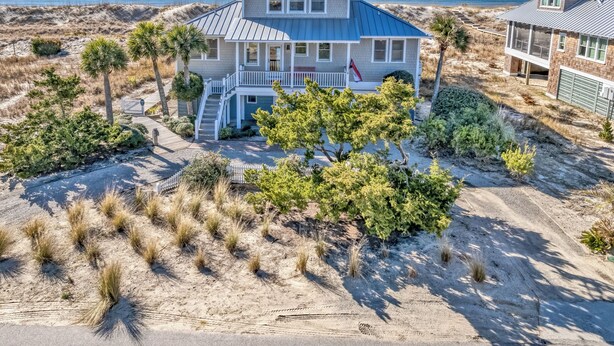 A two-story beachfront house with a covered porch, a flag, and a sandy landscape. - Discover your dream beach house with an amazing ocean view and lots of deck space on South Beach! (Bald Head Island)