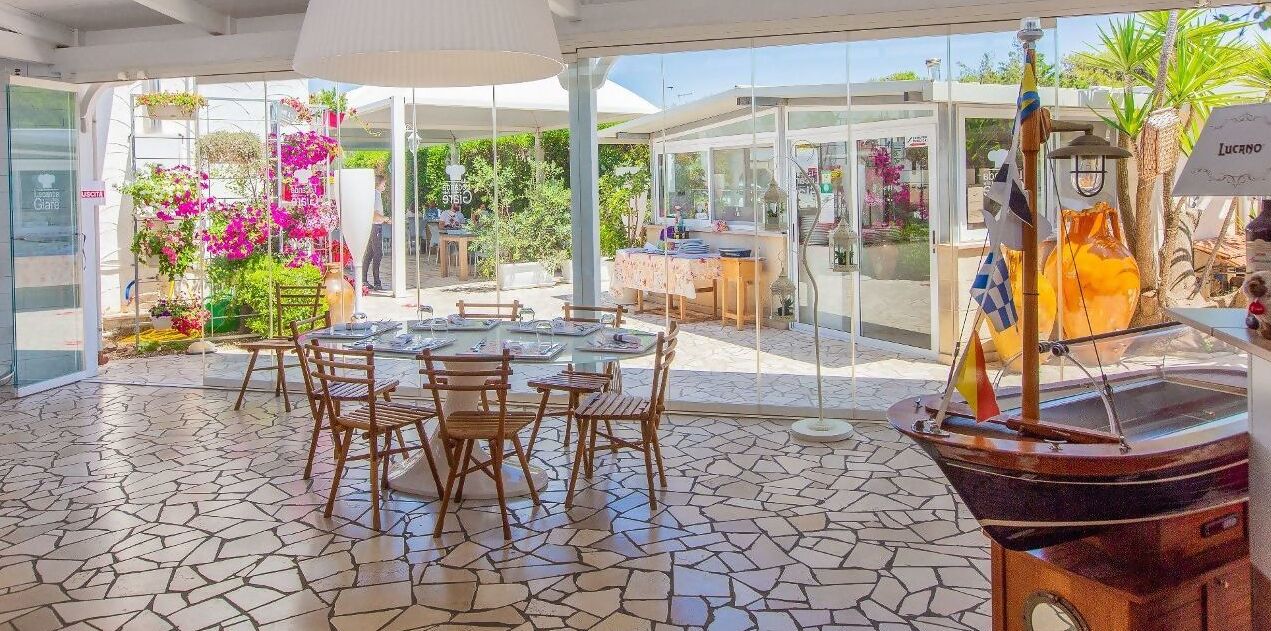 Ampia area patio con tavolo e sedie rotonde, circondata da pareti di vetro che offrono vista su un giardino fiorito e un angolo cucina.