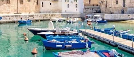 A harbor with boats docked, a stone building with balconies, and a clear blue sky.