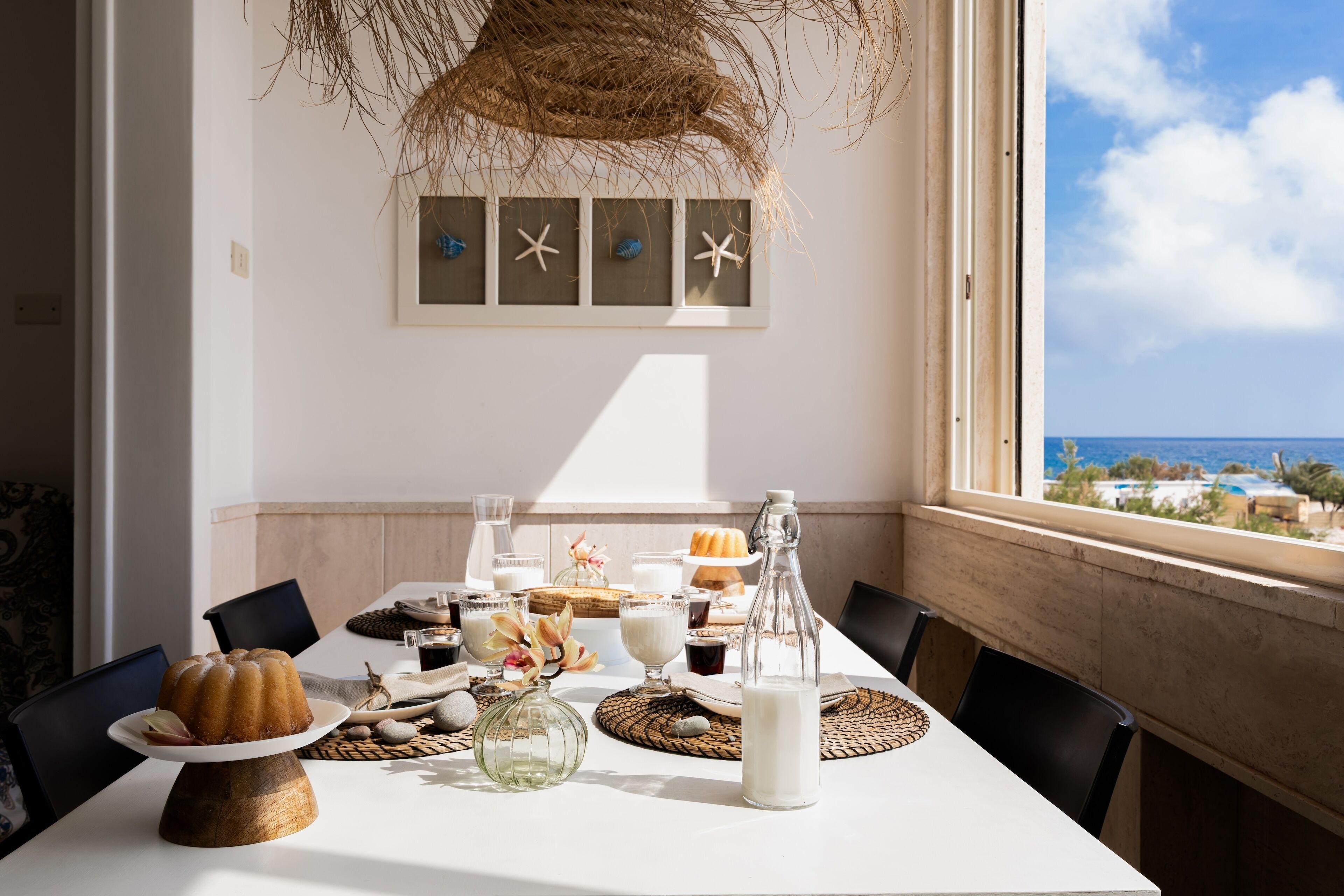 Una mesa de comedor dispuesta con comida y bebidas, vista al océano, y una pared decorada con estrellas de mar.