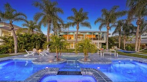 Un área de piscina lujosa con varias zonas de estar, una chimenea central y una casa moderna al fondo.