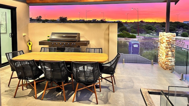 An outdoor dining area with a wooden table and black wicker chairs, a built-in grill, and a view of a sunset over a landscape with trees and a road.