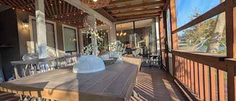 Uma área de refeições ao ar livre coberta, com um deck de madeira, mesa posta com cadeiras e um lustre pendurado.