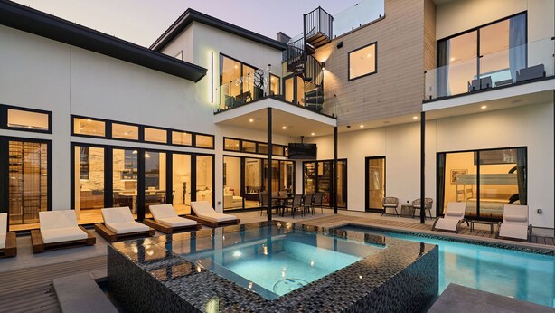 Ein modernes, zweistöckiges Haus mit einem großen Außenpool, mehreren Liegestühlen und einer geräumigen Terrasse.