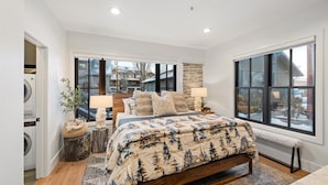 A bedroom with a large bed, two bedside lamps, a stone accent wall, and a view of a snowy landscape through large windows. - The Silver Buffalo Suite in Downtown Whitefish (Whitefish)