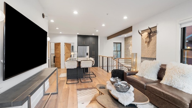 A modern living room with a large flat-screen TV, a sofa, a coffee table, and a dining area with a bar stool. - The Silver Buffalo Suite in Downtown Whitefish (Whitefish)