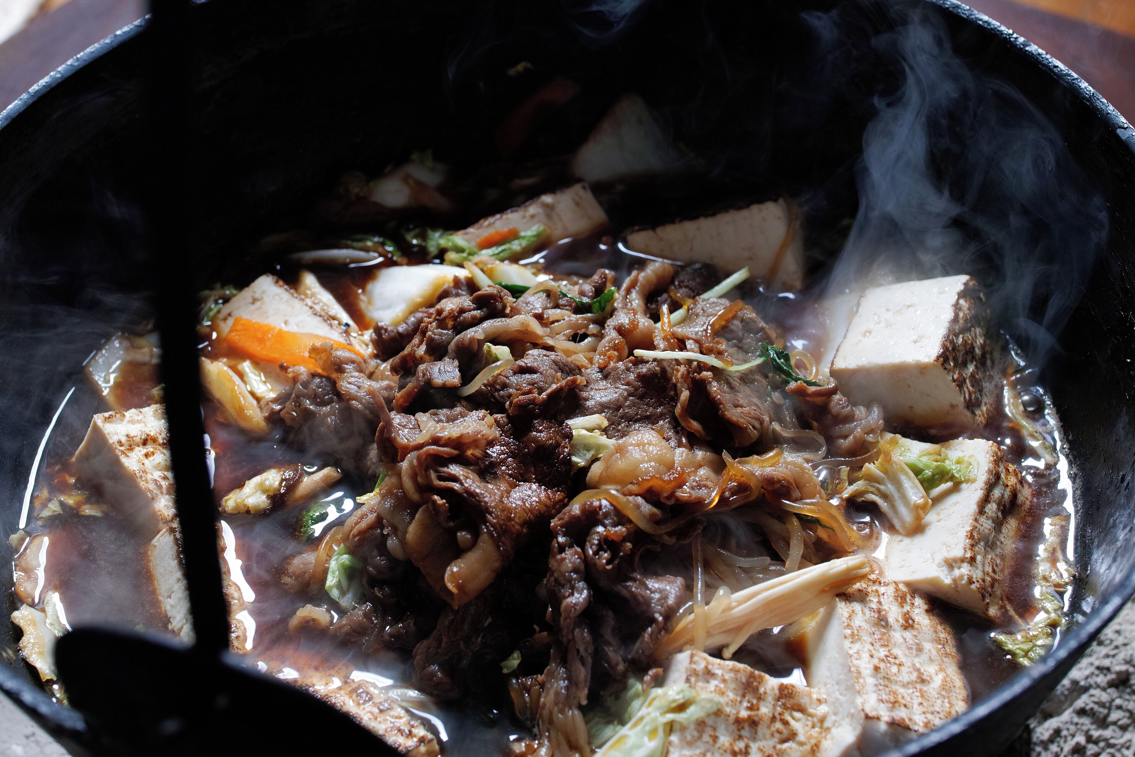 Primer plano de un plato de olla caliente con diversos ingredientes, como carne, tofu y verduras, en una olla humeante.