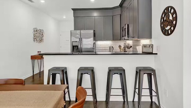 Une cuisine moderne avec des meubles gris, des appareils en acier inoxydable et un plan de travail noir. On y trouve quatre tabourets de bar et une table à manger en bois.