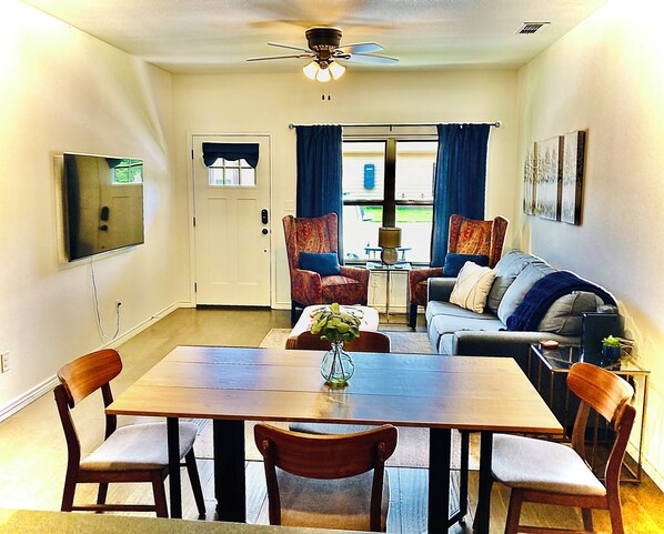 A dining area with a wooden table and chairs, a television mounted on the wall, and a ceiling fan.