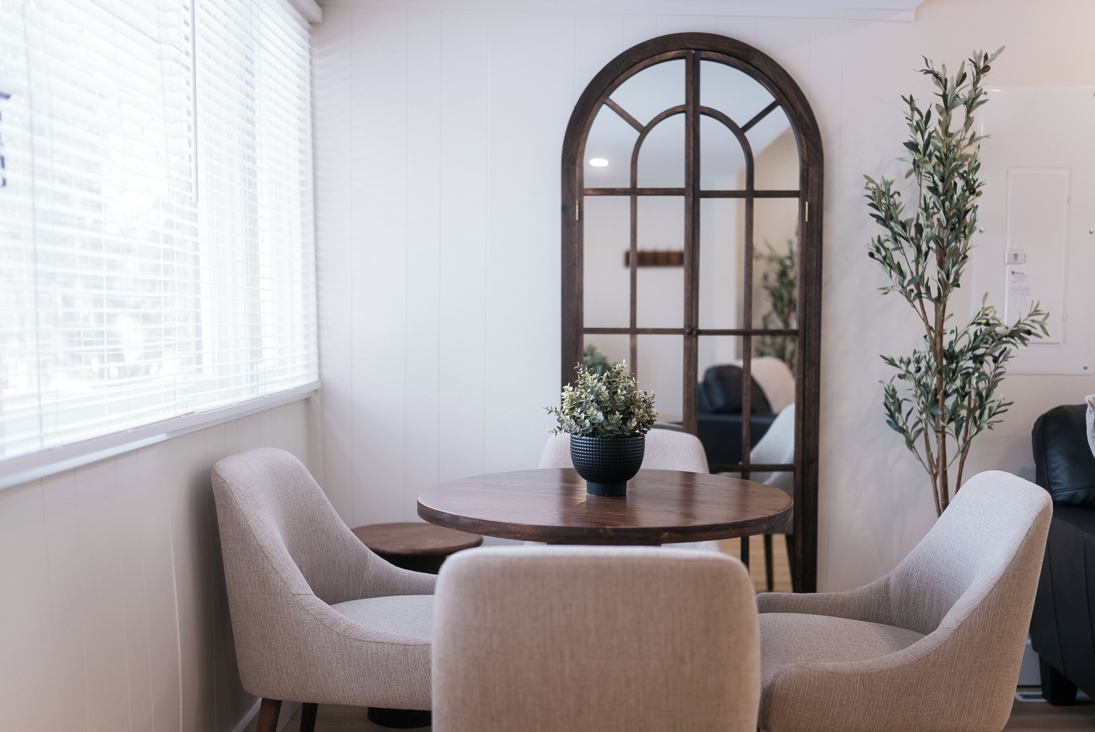 Ein Zimmer mit einem runden Holztisch, zwei beigen Stühlen, einer Topfpflanze und einem großen Bogenförmigen Spiegel.