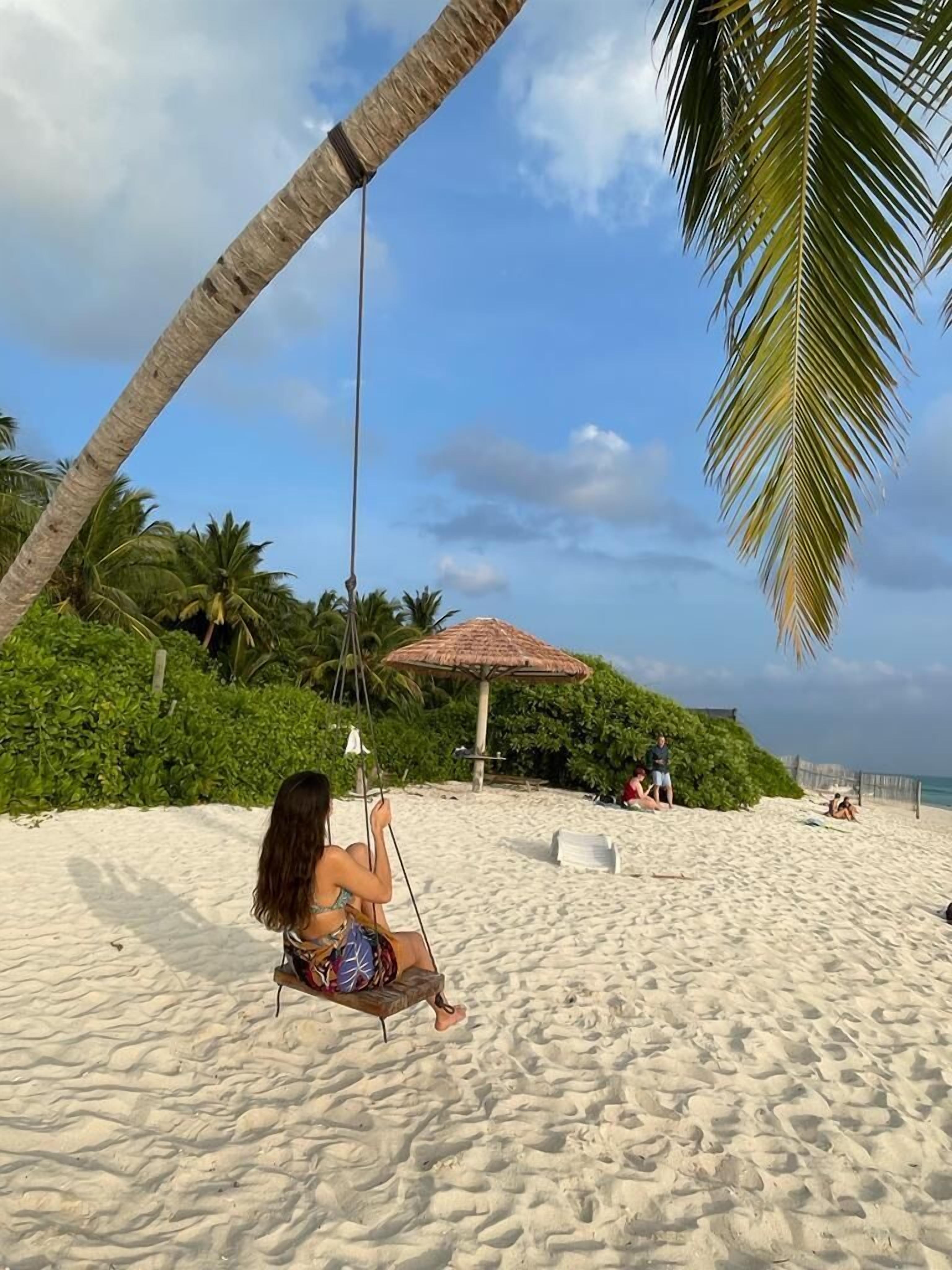 Escena de playa con una silla hamaca bajo una palmera, una sombrilla de paja y gente relajándose en la arena.