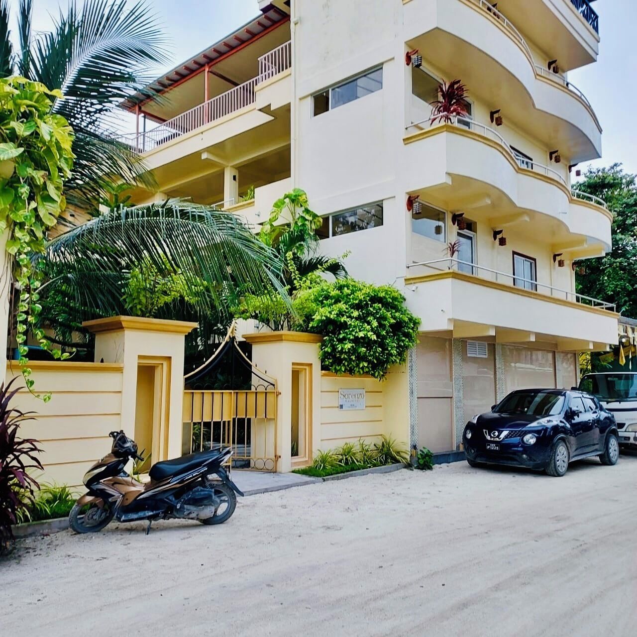 A multi-story residential building with balconies, a gated entrance, and a parked motorcycle and car.