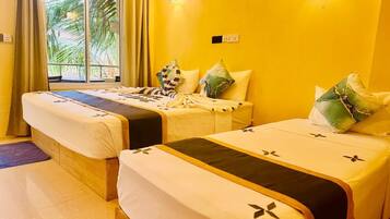 A hotel room with two beds, each with a white bedspread and black trim. There is a window with curtains, a wall-mounted light, and a view of palm trees outside.