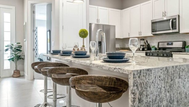 A modern kitchen with white cabinets, stainless steel appliances, and a granite countertop. There are four woven bar stools and a potted plant. - Amazing home in Orlando just 5 minute away from Disney World (Kissimmee)