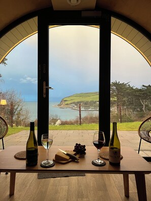 A table with wine bottles and glasses, a cheese board, and a view of the sea through an arched doorway. - Ocean View Pods (Ballymena)