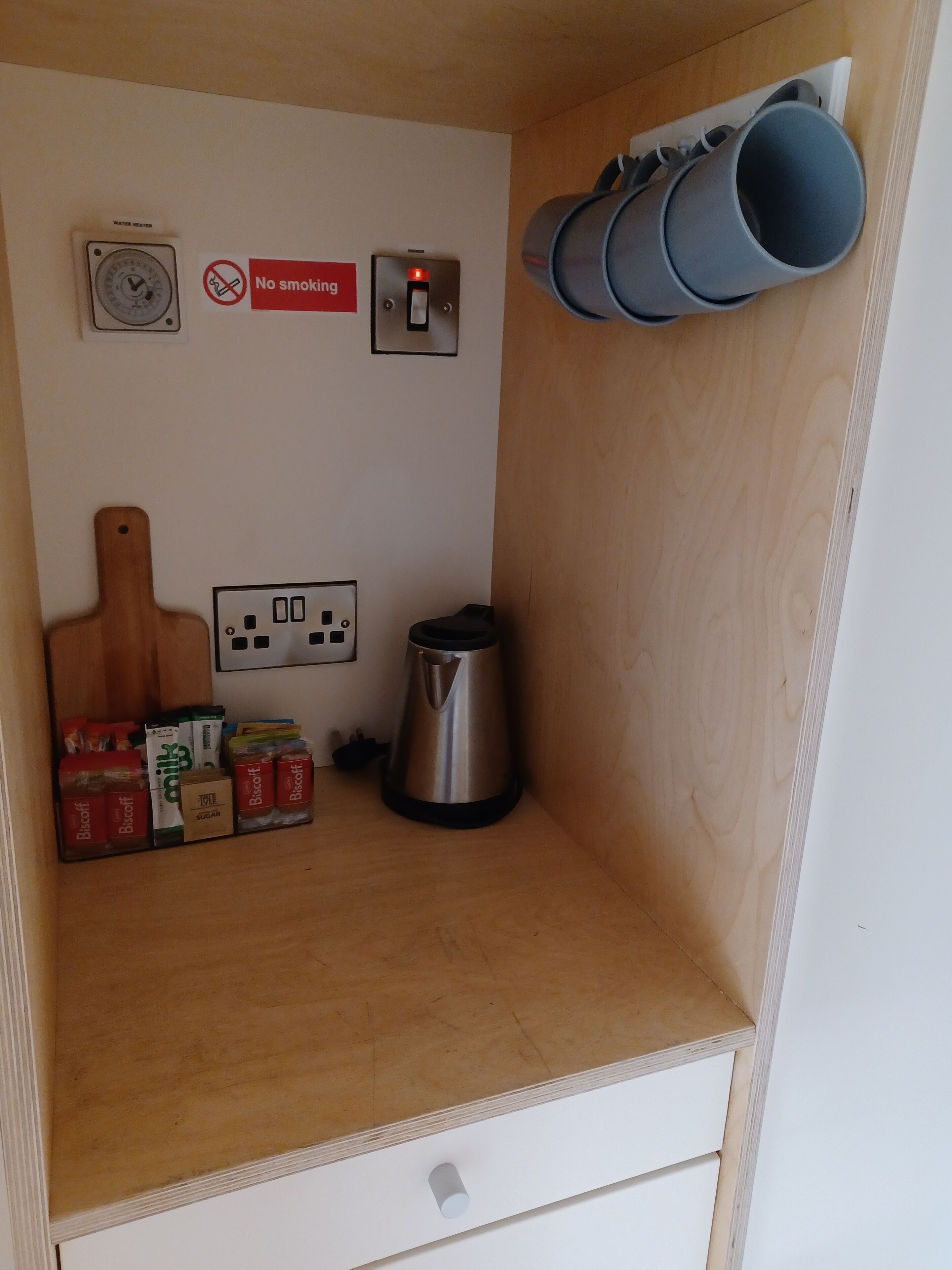 A compact kitchenette with a wall-mounted coffee maker, a no-smoking sign, a power outlet, and a small shelf with kitchen essentials.