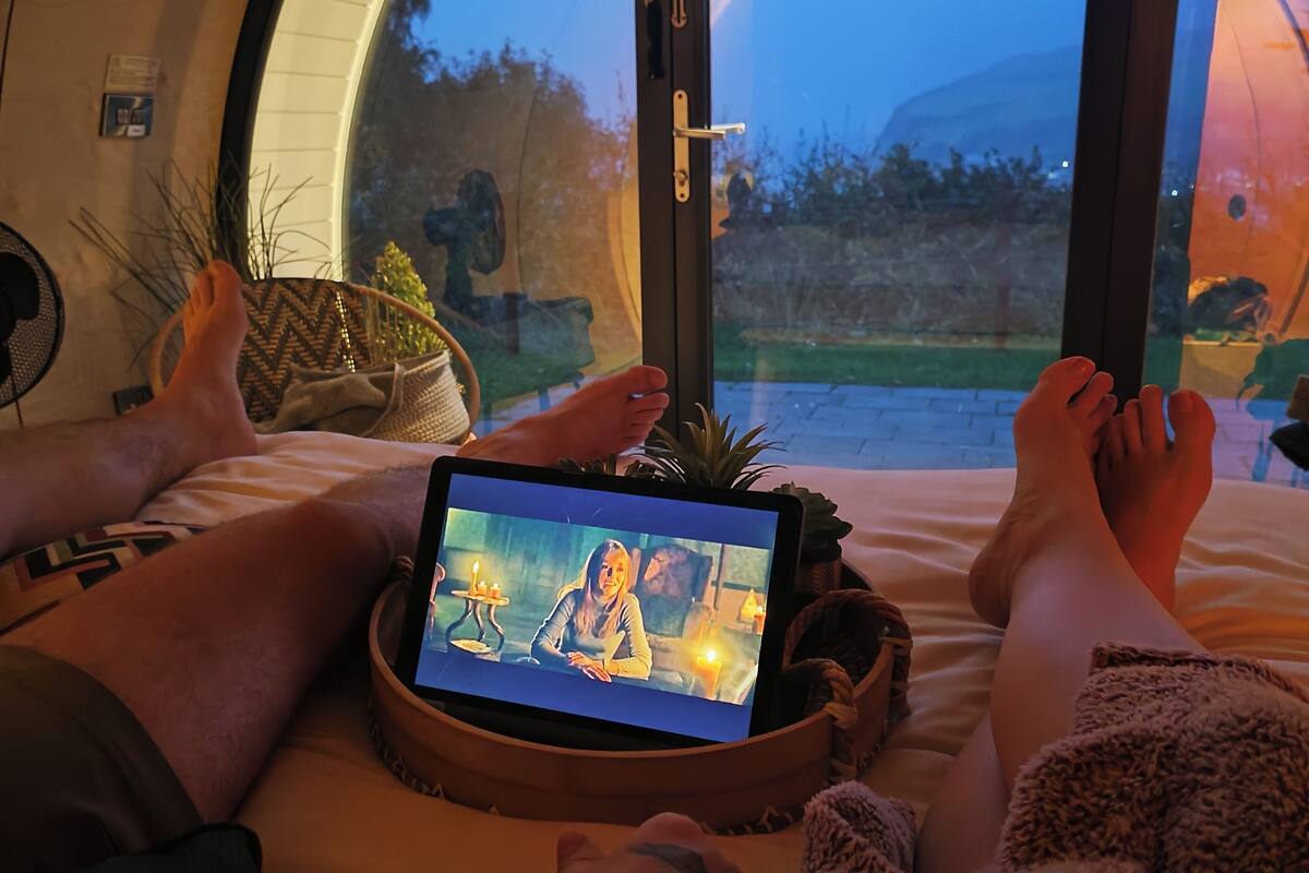 A room with a large arched window, a bed with a tablet displaying a scene, and a cozy blanket.