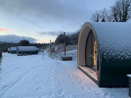 Zona nevada al aire libre con una gran estructura en arco iluminada desde el interior, rodeada de árboles cubiertos de nieve y un camino que lleva a otras construcciones.