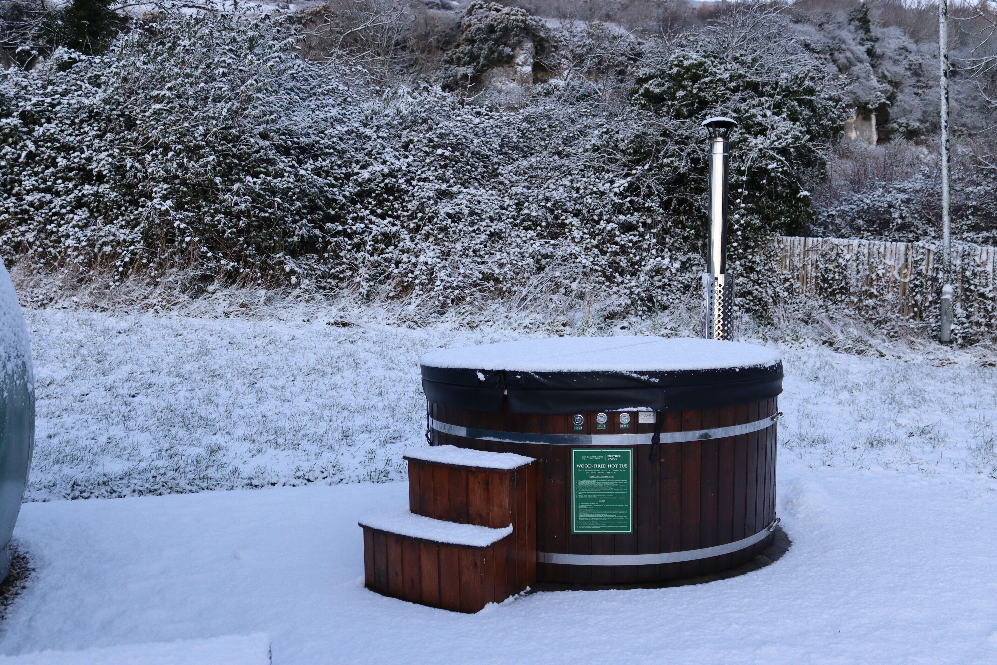 A wooden hot tub surrounded by snow-covered bushes and a wooden fence.