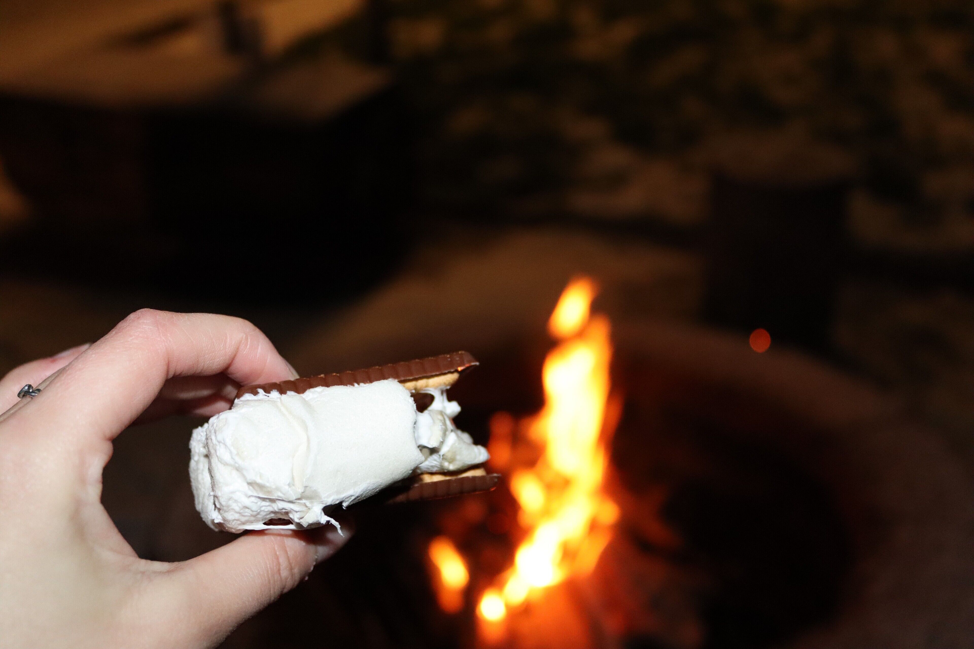 Mão segurando uma barra de chocolate parcialmente comida, com uma fogueira acesa ao fundo.