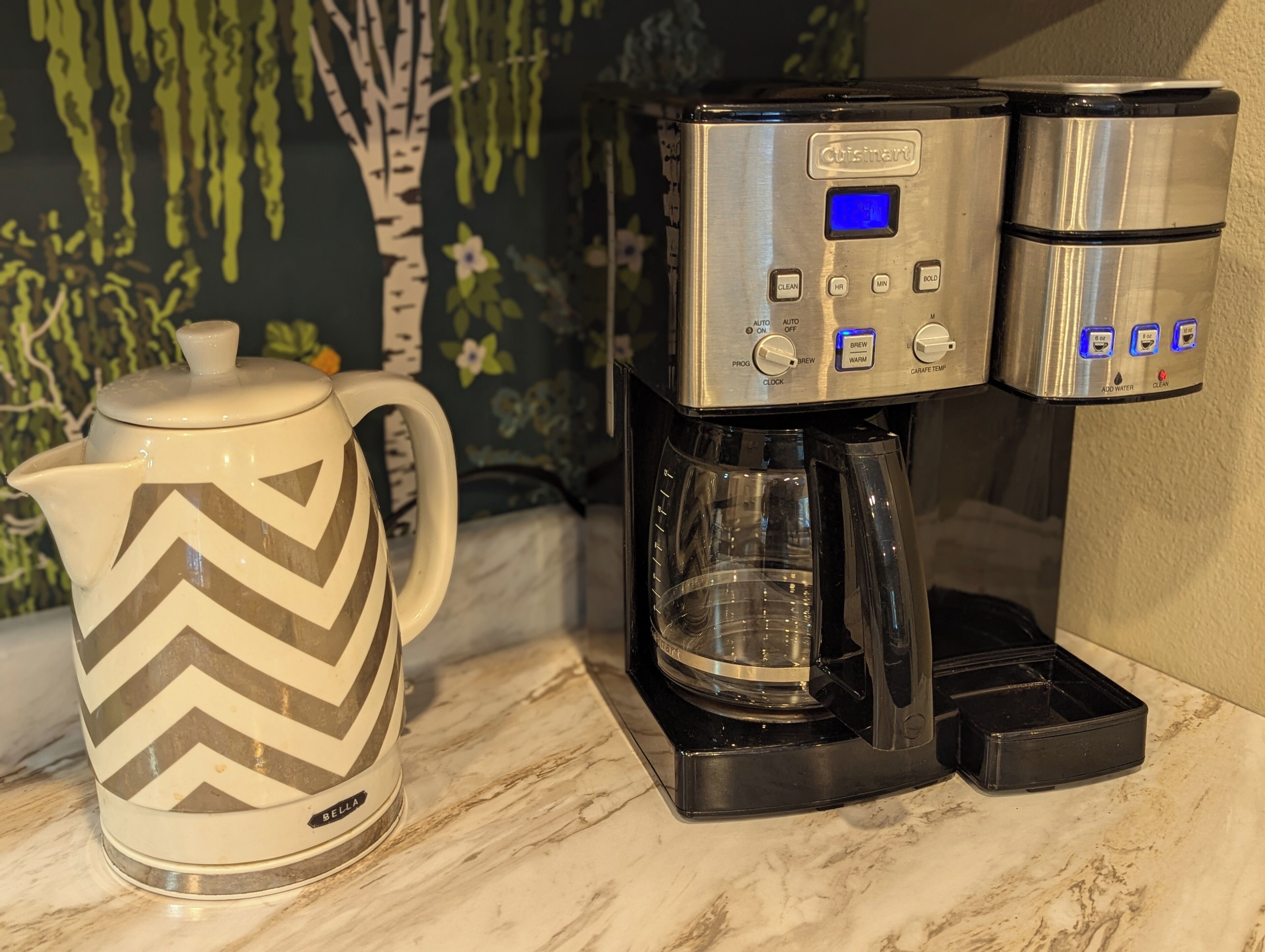 Eine Kaffeemaschine mit digitaler Anzeige und Bedienelementen, einer transparenten Glascarafe und einer weißen Tasse mit Zickzackmuster.