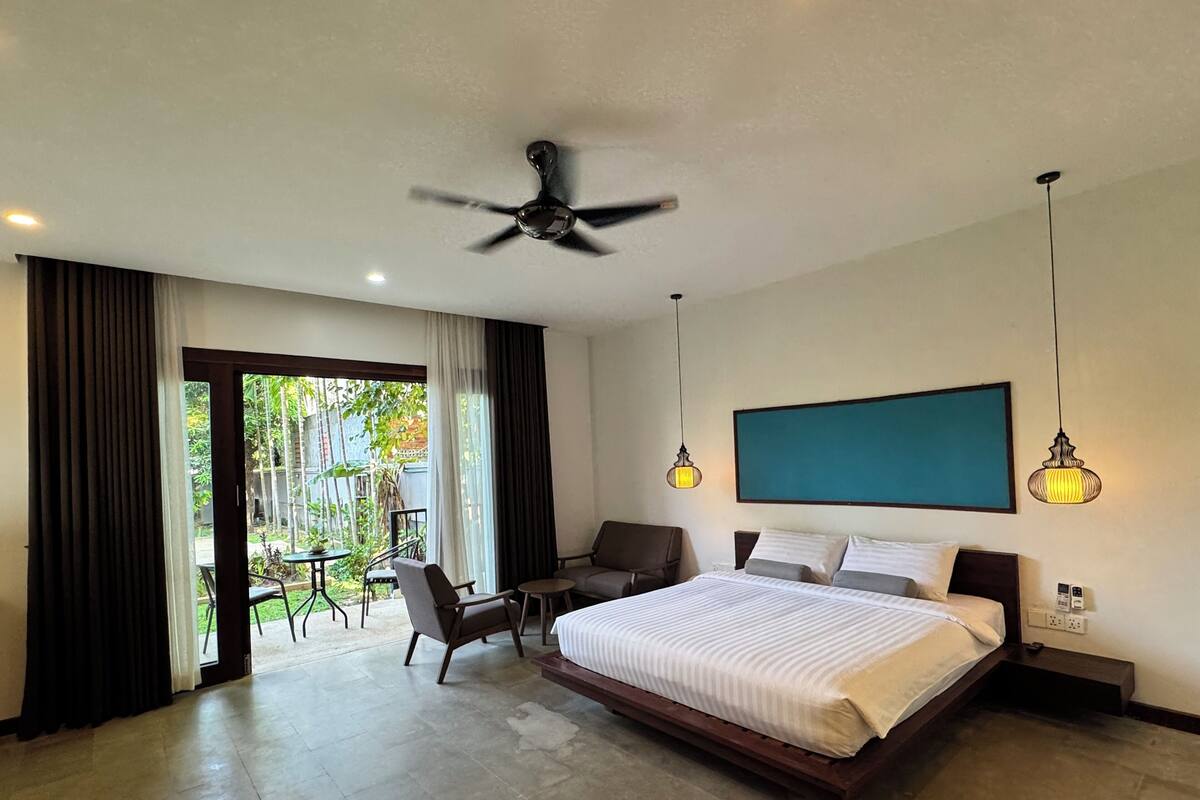 Una habitación de hotel moderna con una cama grande, un ventilador de techo, una mesa de comedor con sillas y vistas al exterior.