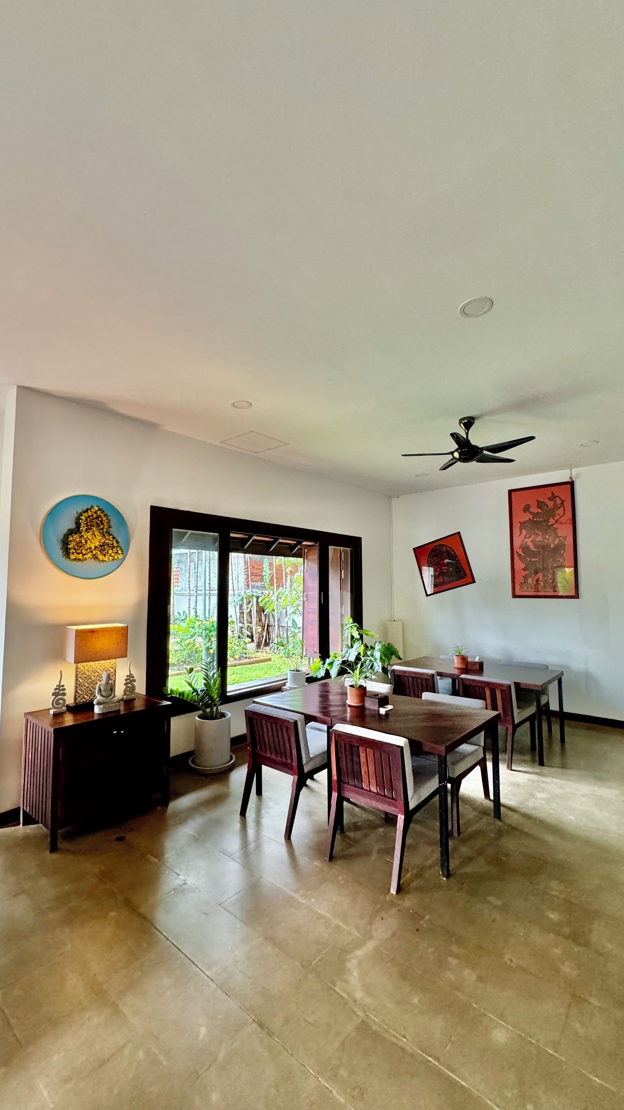 Uma ampla área de jantar com uma grande mesa e cadeiras, um ventilador de teto e vista para um jardim exterior através de uma porta de vidro.