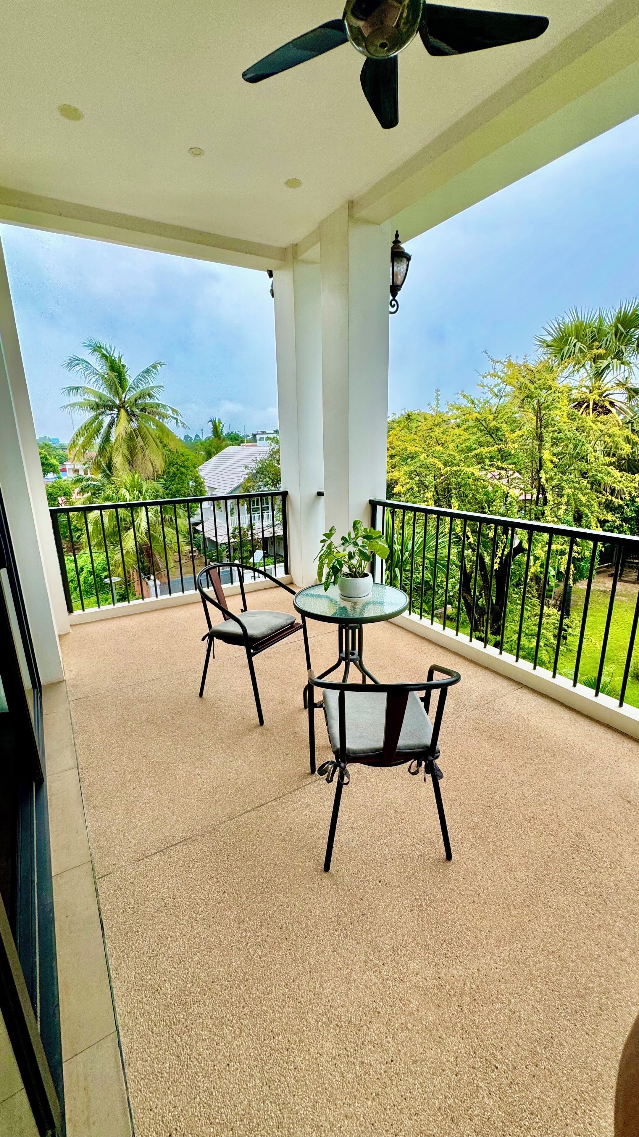 A balcony with a ceiling fan, two chairs, a small table with a potted plant, and a view of greenery and buildings.