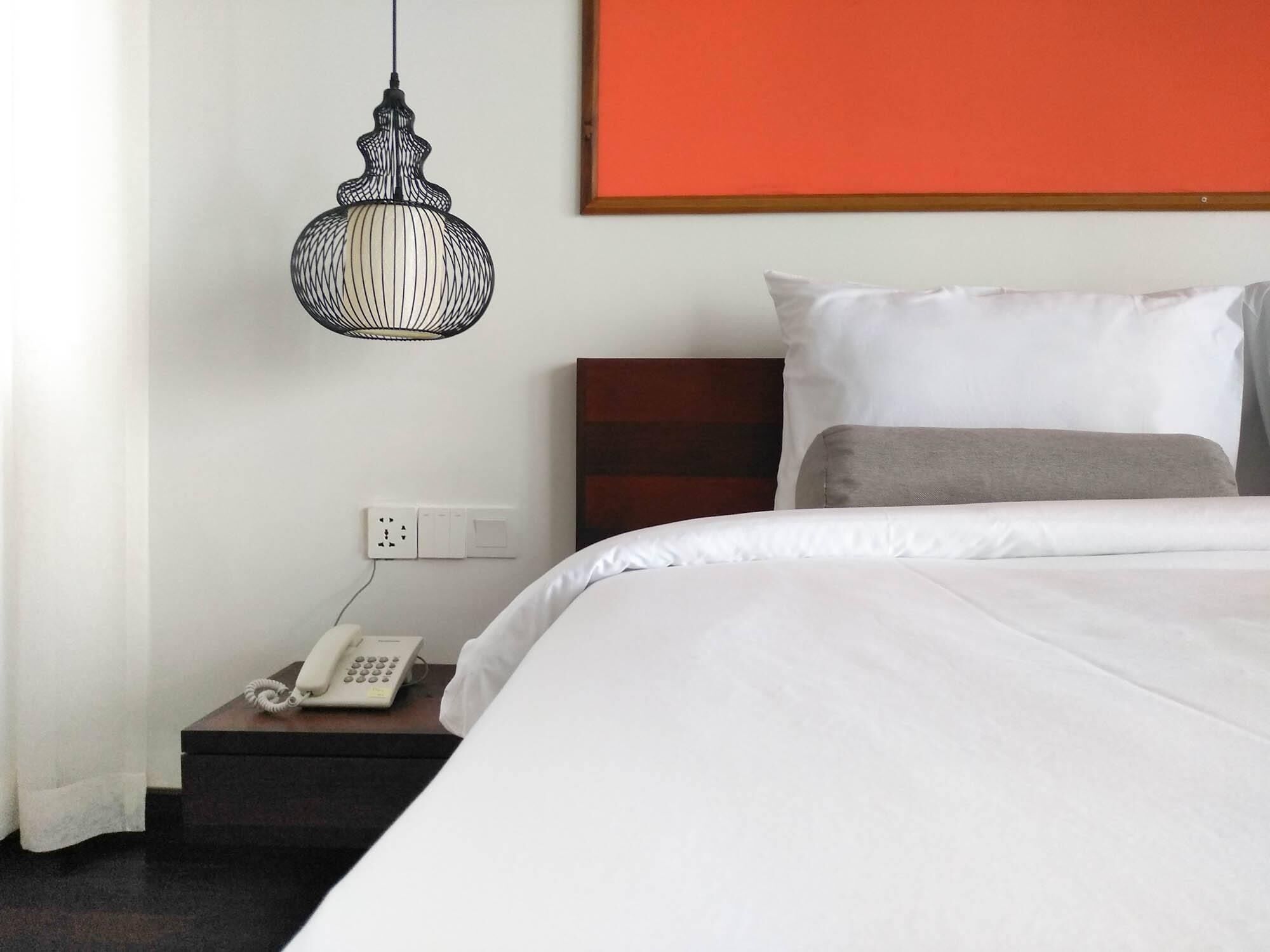 A hotel room with a bed, a bedside telephone, a power outlet, and a hanging light fixture.