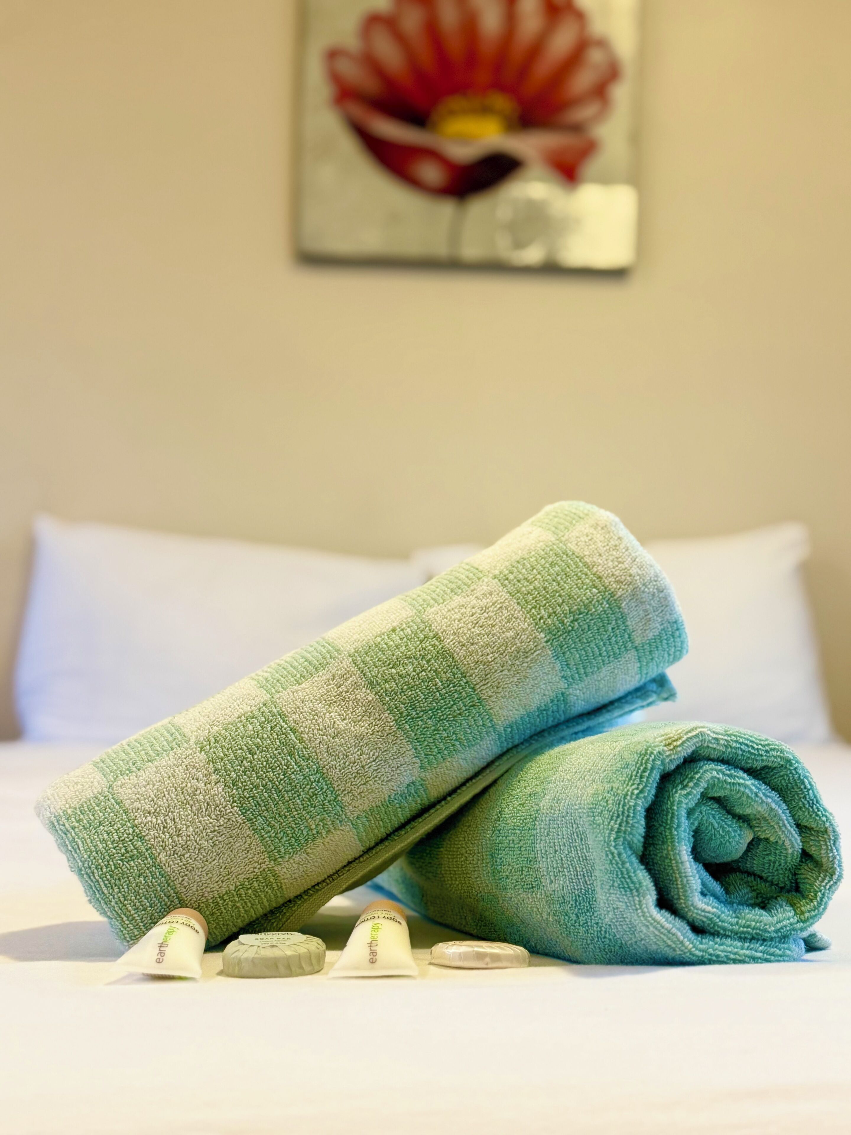 A neatly made bed with a green and white checkered towel, two small bottles labeled 'earththerapy', and a painting of a flower on the wall.