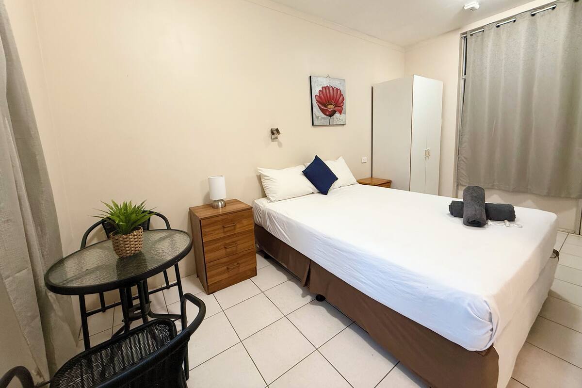 A bedroom with a bed, bedside table, lamp, and a small table with a plant.