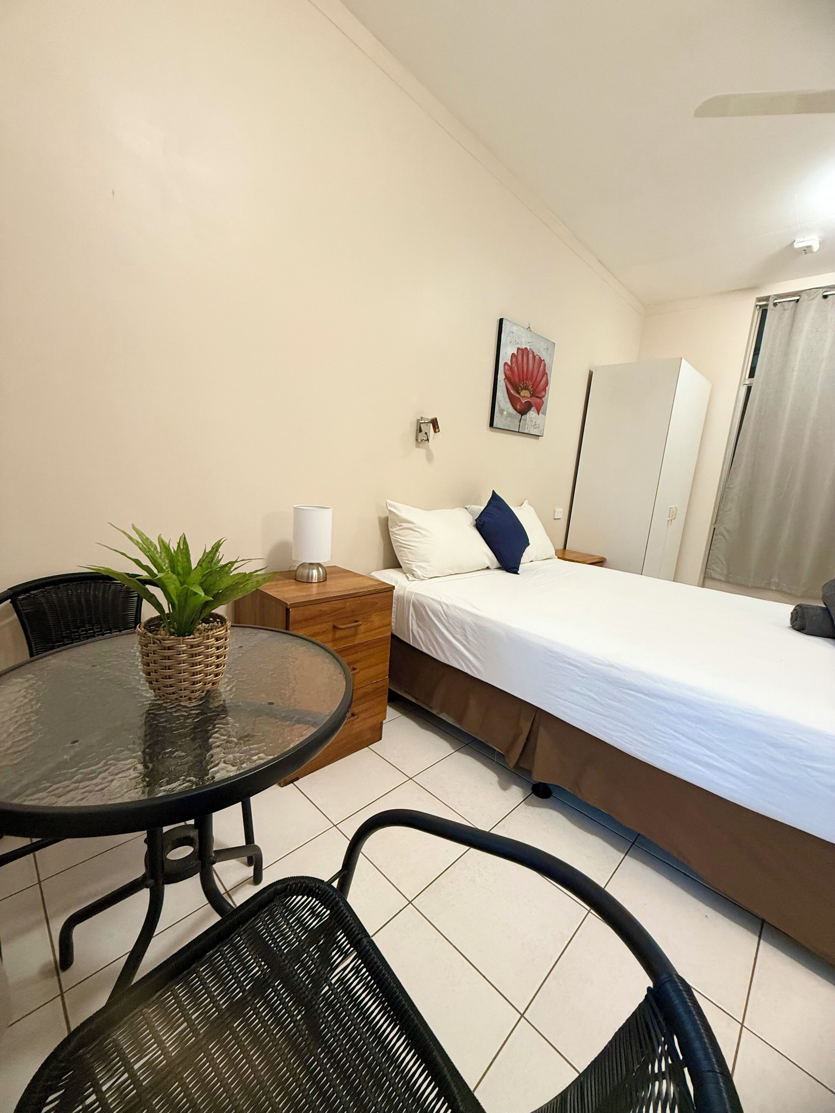 A hotel room with a bed, a small table with a plant, a chair, a nightstand, and a wall-mounted lamp.
