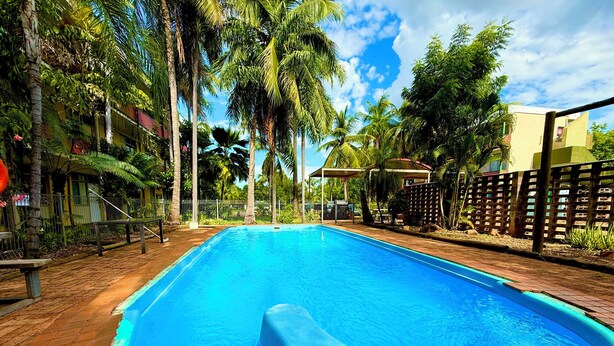 A rectangular swimming pool with clear blue water, surrounded by a brick walkway and palm trees. - Tropical River Hotel (Katherine)
