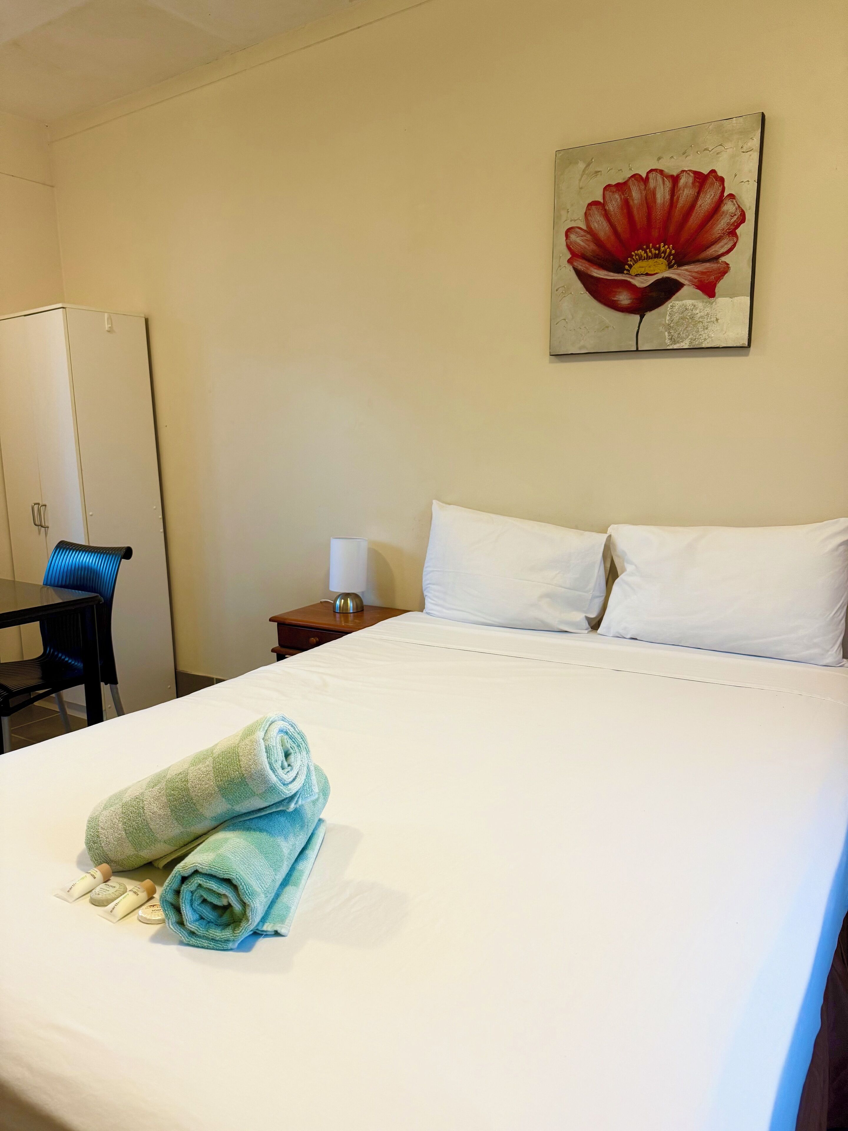 A hotel room with a bed, a desk, a chair, a nightstand, a lamp, and a painting of a flower on the wall.