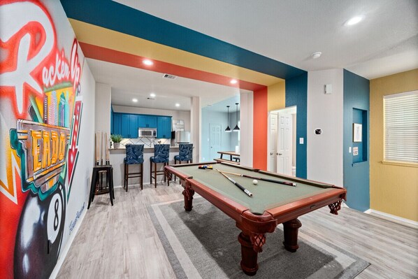 Une table de billard dans une pièce ornée d’une fresque murale colorée, avec un coin cuisine et un bar doté de chaises bleues.