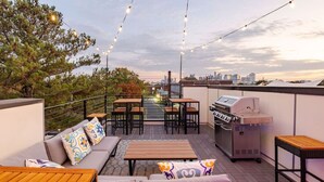 An outdoor patio with a grill, seating area, and string lights against a city skyline.