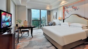 A hotel room with a large bed, a desk with a TV, a chair, a small table, and a view of the city.